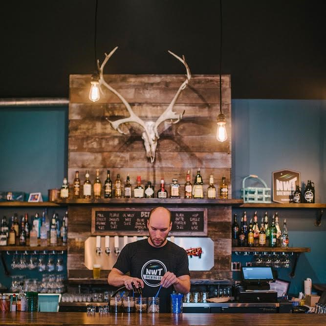 A male bartender stands behind a counter mixing drinks, behind him are an array of bottles and a large skull of a moose mounted to a wooden board 