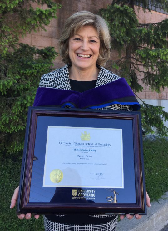 Profile of Shirlee Sharkey standing with her honorary doctor of laws degree framed in front of her