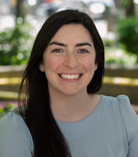 The image shows a young woman smiling. She has long brown hair, green eyes, and is wearing a light blue top.