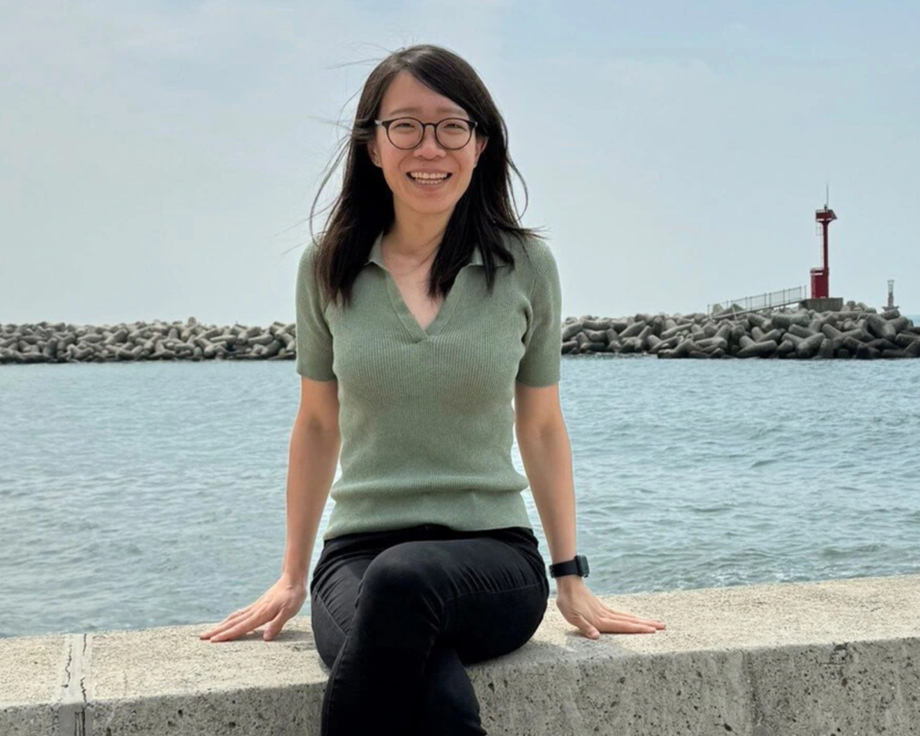 A woman with long black hair and glasses smiles while sitting outdoors on a concrete ledge by the water. She is wearing a light green short-sleeved top and black pants. Behind her, there is a calm body of water, a rocky breakwater, and a red lighthouse in the distance.