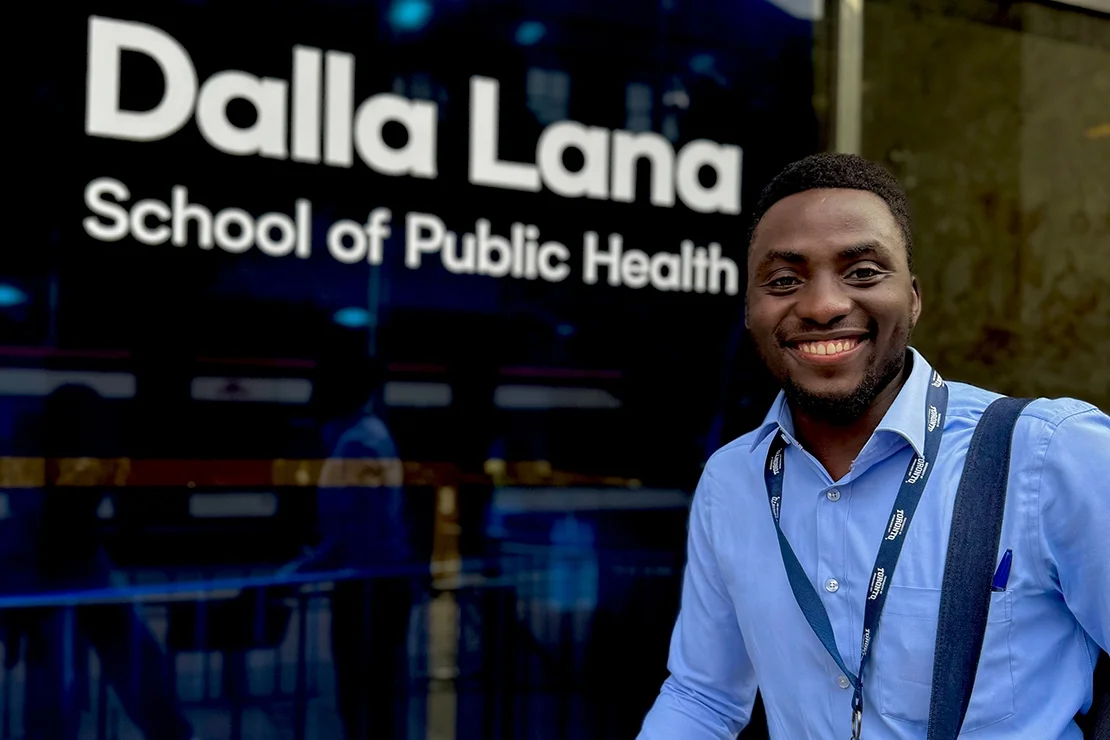 A smiling individual stands in front of a building with the signage 'Dalla Lana School of Public Health' visible in the background. The person is wearing a light blue button-up shirt and a lanyard around their neck.