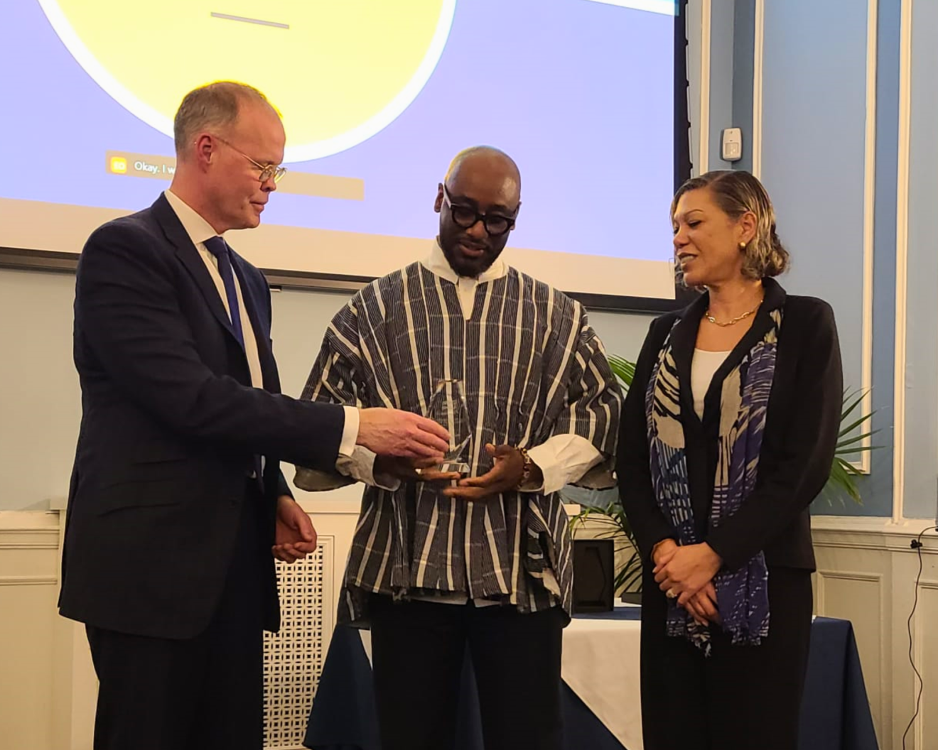 Three individuals stand on stage, one in the center holding an award. The recipient, wearing a traditional patterned garment, is presented the award by a man in a suit on the left, while a woman in business attire on the right looks on with a smile. The background displays a screen with a presentation. Health Administration Student Advancing Equity.