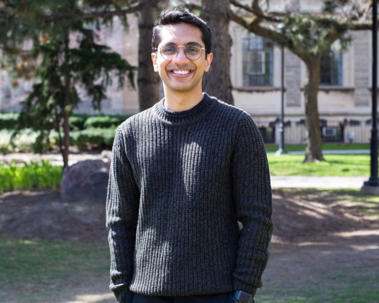 A young man smiling happily outside on a sunny day with academic buildings seen in the background. Vinyas Harish reflects on his time in the CEHCR program.