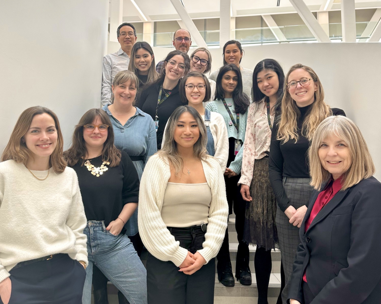 A group photo of a diverse team from the Women’s Age Lab, gathered on a staircase in a bright, modern building. The group includes individuals of varying ages and backgrounds, smiling warmly for the camera. Natural light illuminates the space, highlighting their professional yet friendly demeanor. The members of the Women's Age Lab.