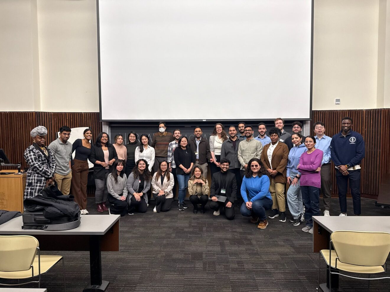 A group of people are posing for a photo in a classroom during the AI Hackathon. They are standing and kneeling in front of a large white projection screen. The room has wooden paneling on the walls and several desks with chairs arranged in rows. Some individuals are wearing masks, and various casual outfits can be seen.