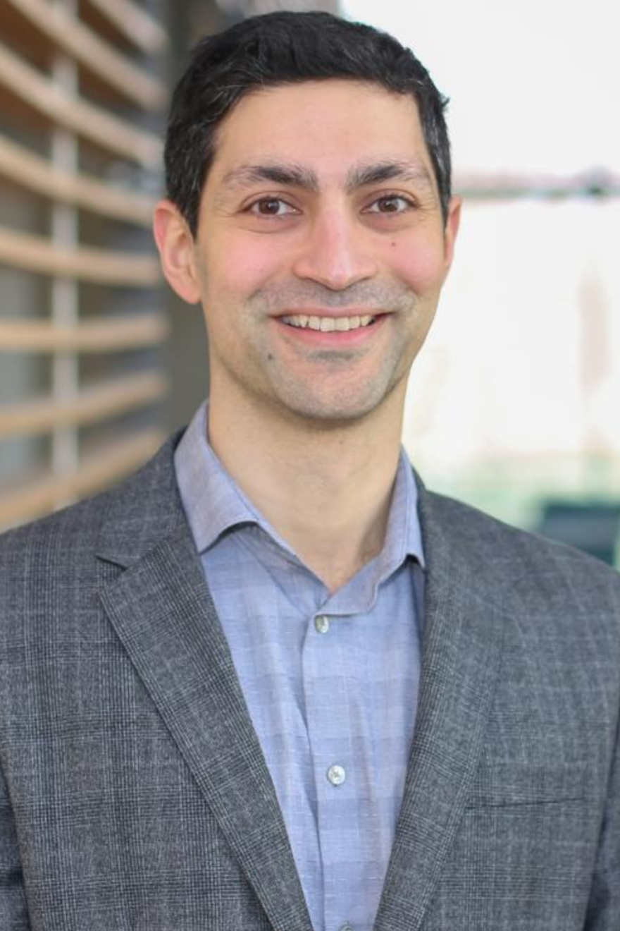 A person, a 2025–26 Vector Scholar, wearing a grey blazer and light blue button-up shirt stands in front of a blurred background with horizontal wooden slats on the left.