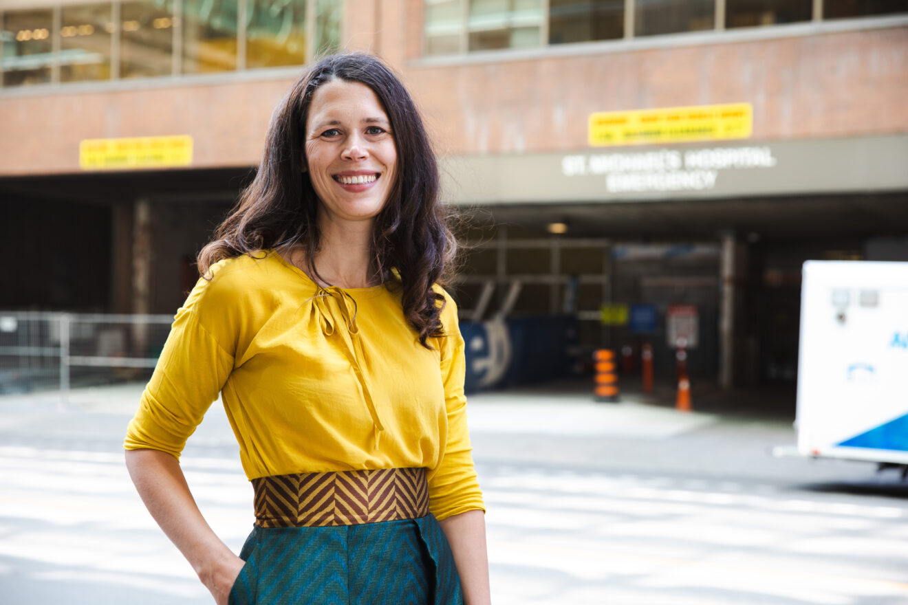 An image of Dr. Tara Gomes, lead of a study on safer opioid supply, wearing a light-colored blouse and a patterned skirt. appears to be an outdoor urban setting with buildings, a hospital, and signs, partially blurred.